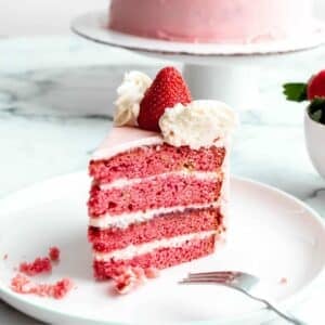 A slice of strawberry cake with cream cheese frosting sitting on a plate, showing the layers of fluffy pink cake and icing.