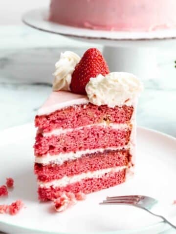 A slice of strawberry cake with cream cheese frosting sitting on a plate, showing the layers of fluffy pink cake and icing.