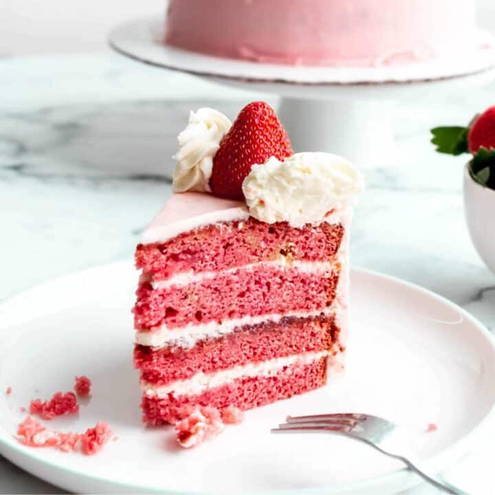 A slice of strawberry cake with cream cheese frosting sitting on a plate, showing the layers of fluffy pink cake and icing.