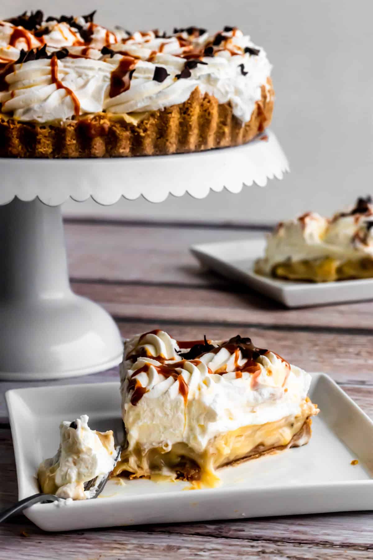 Slice of banoffee pie on a plate with a bite taken out to show the gooey caramel filling, with the whole pie on a cake stand in the background.