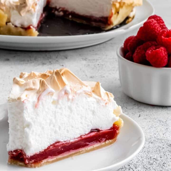 A slice of raspberry meringue pie showing the raspberry curd, and fluffy meringue on a buttery pastry crust.