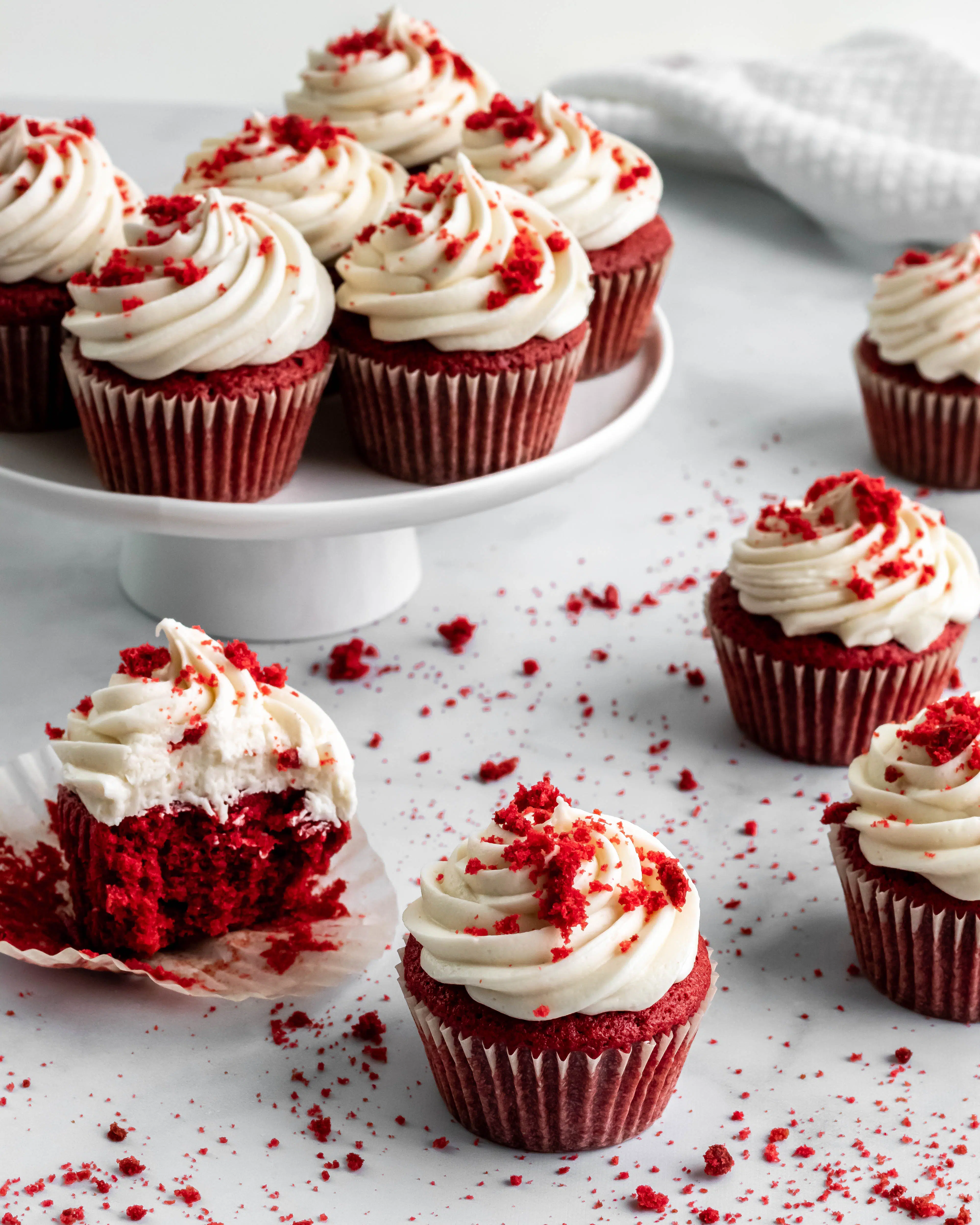 Red Velvet Cupcakes scattered on a table, some are on a cake stand and some are on the table, one has a bite taken out of it. 