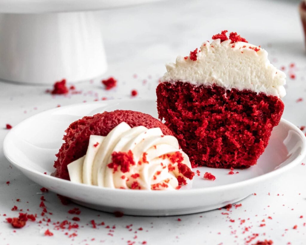 A red velvet cupcake on a plate, sliced in half to show the velvety texture of the crumb inside. 