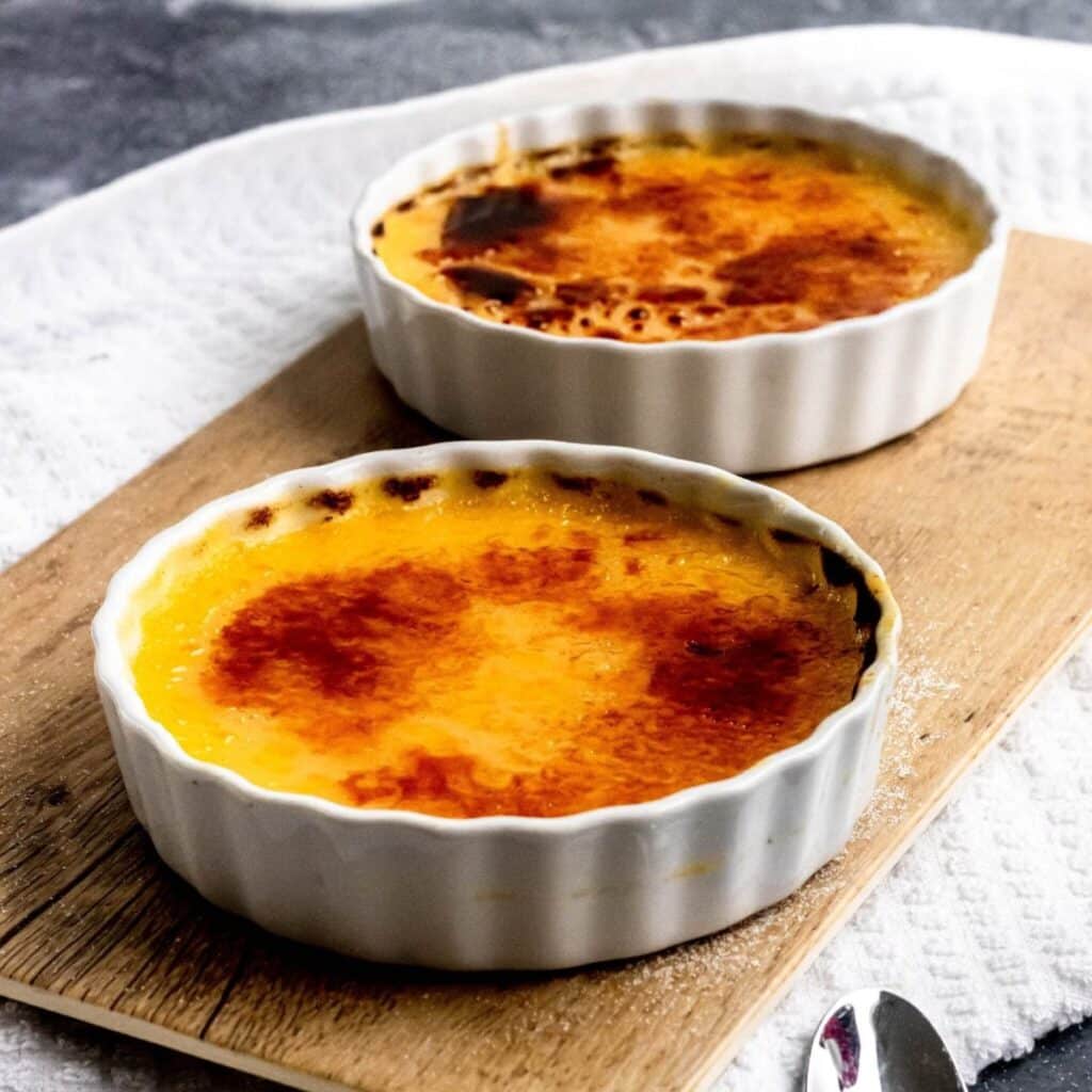 Two crème brulee in round ramekins sitting on a cutting board.