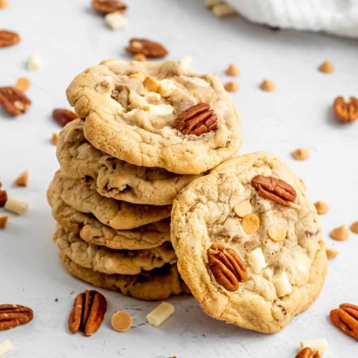 Six white chocolate pecan cookies stacked up, with another one leaning against the stack, showing the top side of the cookie to the camera.