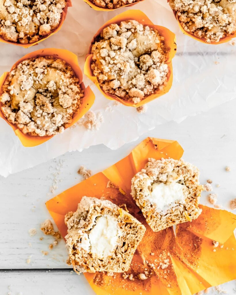 A view from above of cheesecake stuffed carrot muffin cut in half, cut side facing up, and another two standing upright, showing the crumble topping. 