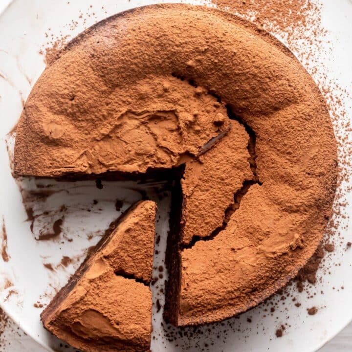Thick chocolate torte, with a sliced taken out of it, on a plate, dusted heavily with coco powder.