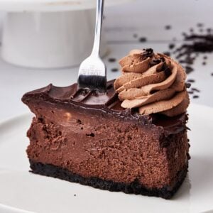 Slice of chocolate cheesecake with a fork sticking out the top showing the thick, creamy texture, with chocolate ganache topping and a swirl of chocolate whipped cream on top.