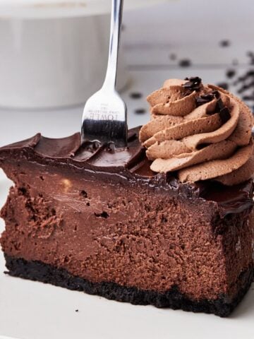 Slice of chocolate cheesecake with a fork sticking out the top showing the thick, creamy texture, with chocolate ganache topping and a swirl of chocolate whipped cream on top.