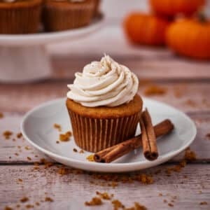 A single pumpkin spice cupcake with a swirl of cream cheese frosting on a plate with two cinnamon sticks next to it.