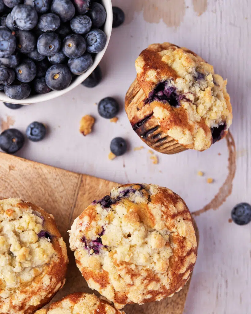 Muffins on a wooden platter with one muffin fallen off, bowl of blueberries next to them.