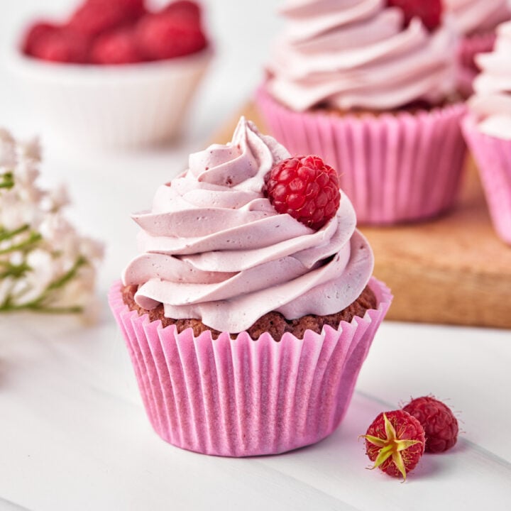 Side view of a finished raspberry cupcake with a swirl of raspberry buttercream and a single raspberry placed slightly off center of the peak of the swirl.