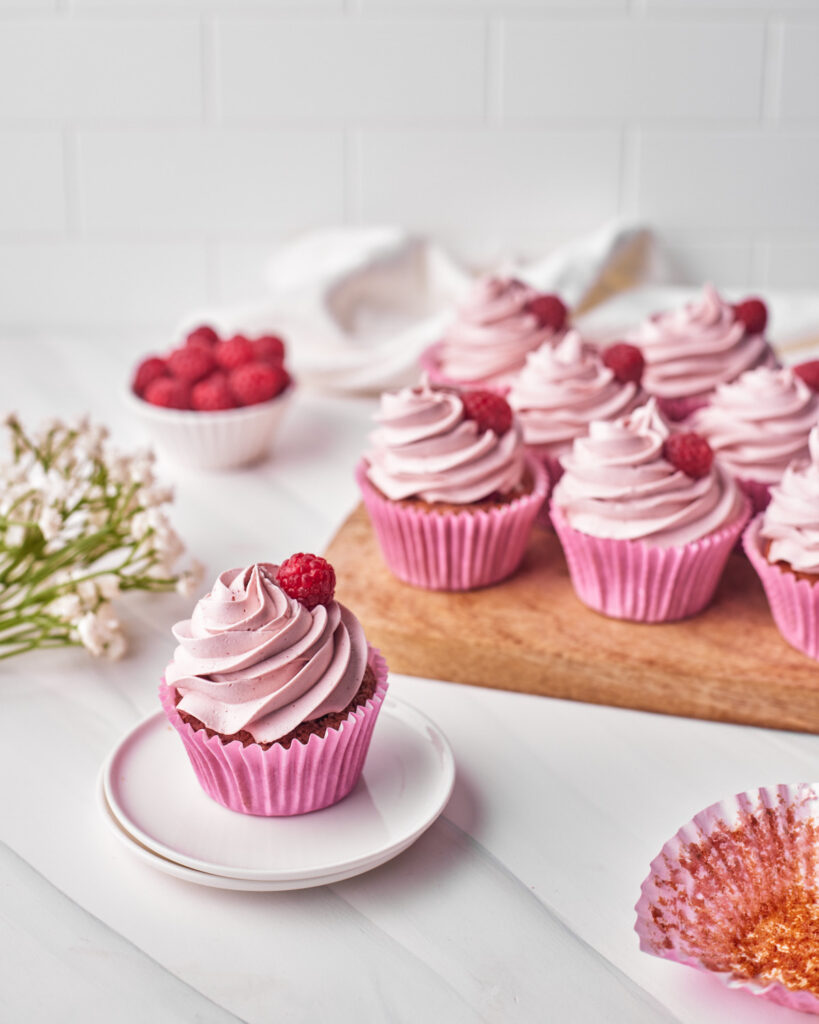 raspberry cupcakes all in a row on a chopping board with raspberry buttercream and a fresh raspberry on top.
