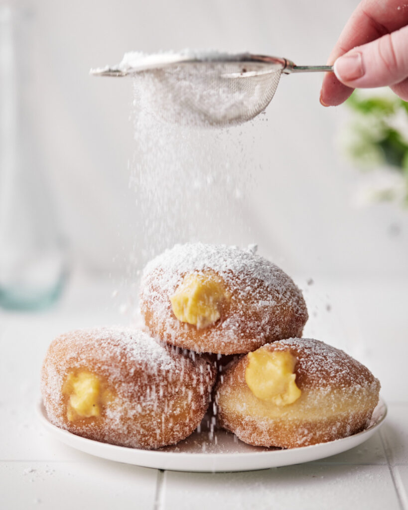 powdered sugar being sprinkled on top of custard donuts. 