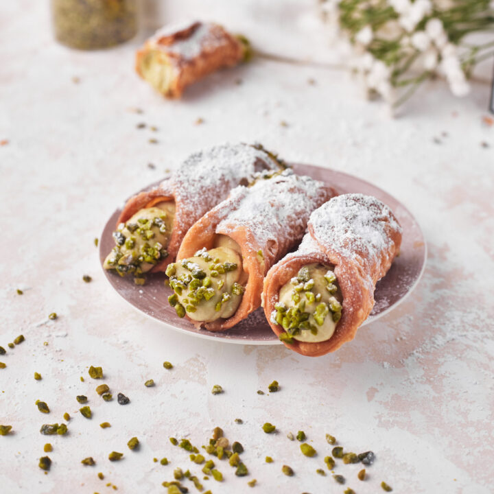 three pistachio cannoli on a plate with the ends dipped in chopped pistachios.
