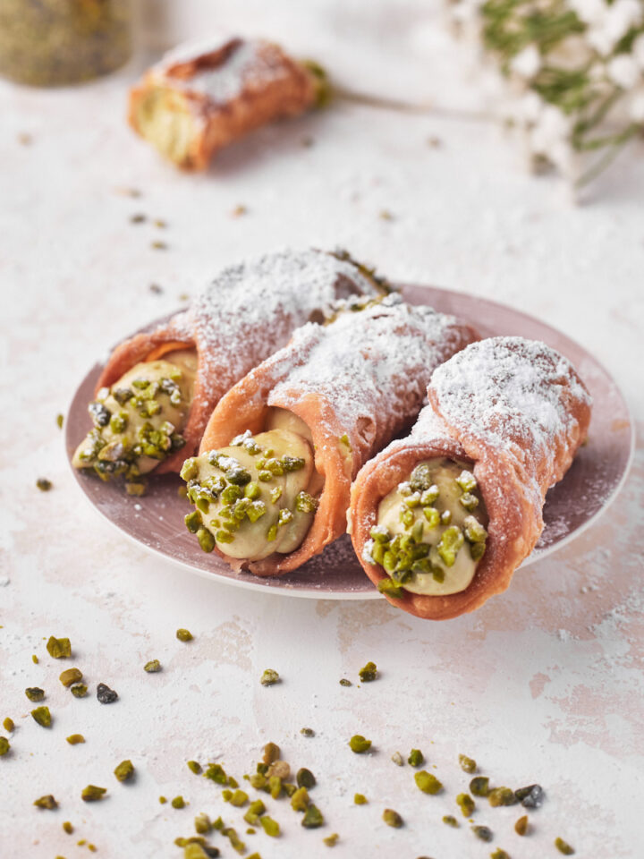 three pistachio cannoli on a plate with the ends dipped in chopped pistachios.
