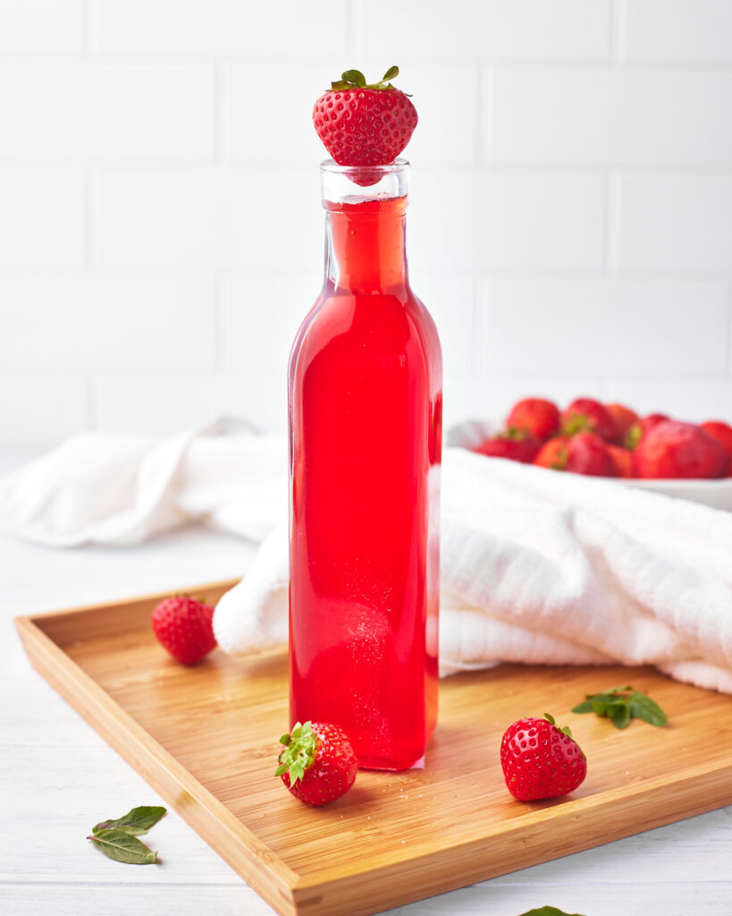 bottle of homemade strawberry simple syrup on a wooden board, surrounded by fresh strawberries.