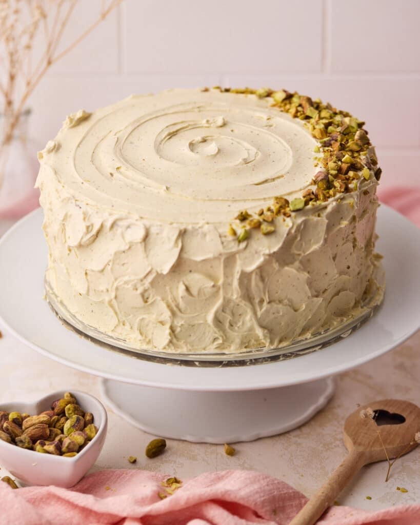 Pistachio cake on a cake stand, covered with soft pistachio buttercream and chopped pistachios.