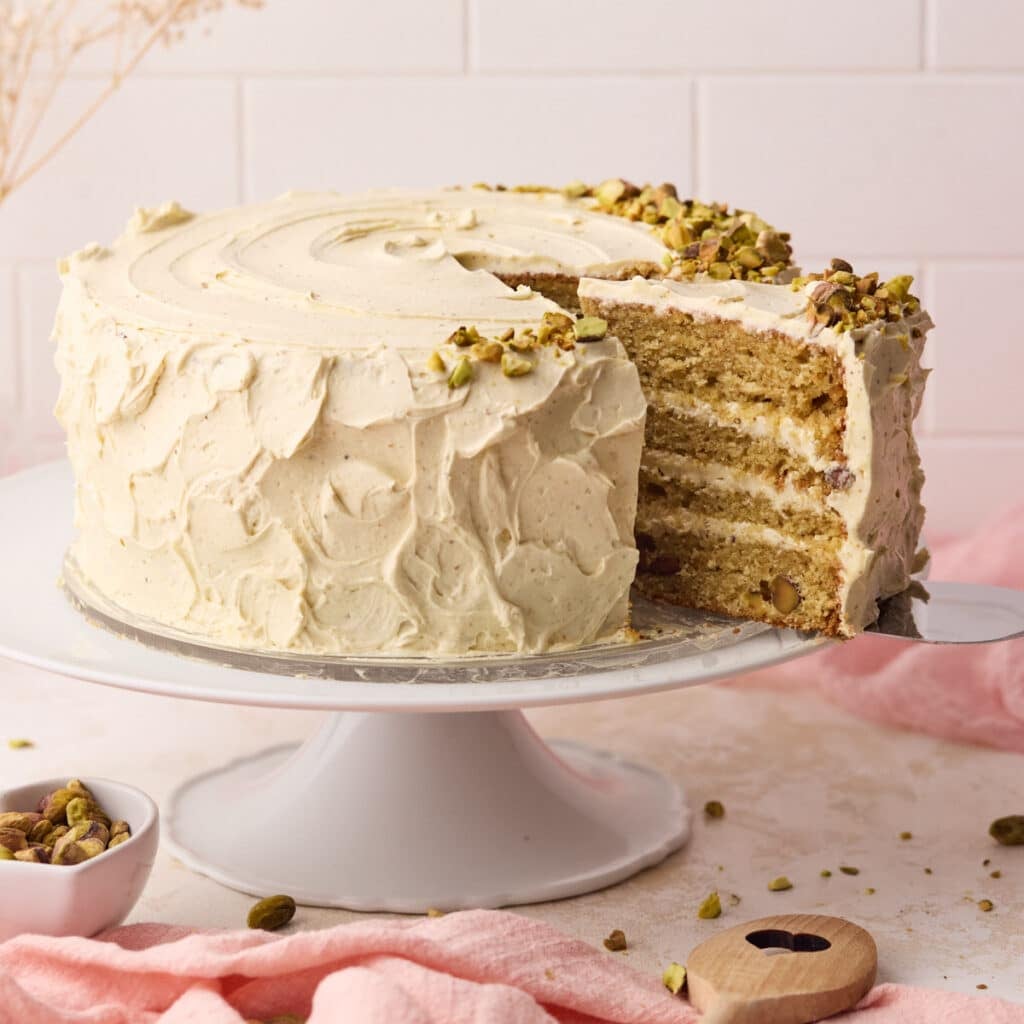 Pistachio cake on a cake stand with a slice being taken out of it to reveal the layers of fluffy cake and italian pistachio buttercream inside.
