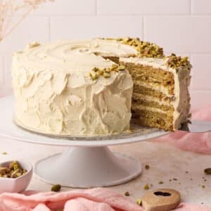 Pistachio cake on a cake stand with a slice being taken out of it to reveal the layers of fluffy cake and italian pistachio buttercream inside.