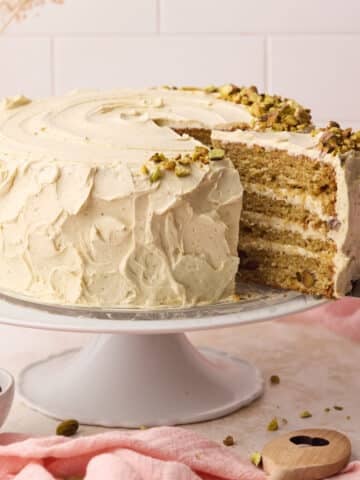 Pistachio cake on a cake stand with a slice being taken out of it to reveal the layers of fluffy cake and italian pistachio buttercream inside.