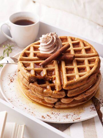 stack of cinnamon waffles on a plate with a swirl of cinnamon whipped cream, served on a breakfast tray in bed.