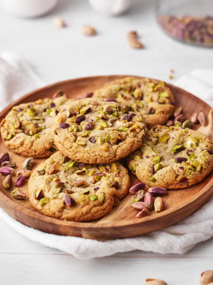 pile of pistachio cookies on a wooden platter, surrounded by pistachio nuts.