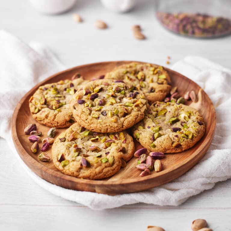 pile of pistachio cookies on a wooden platter, surrounded by pistachio nuts.
