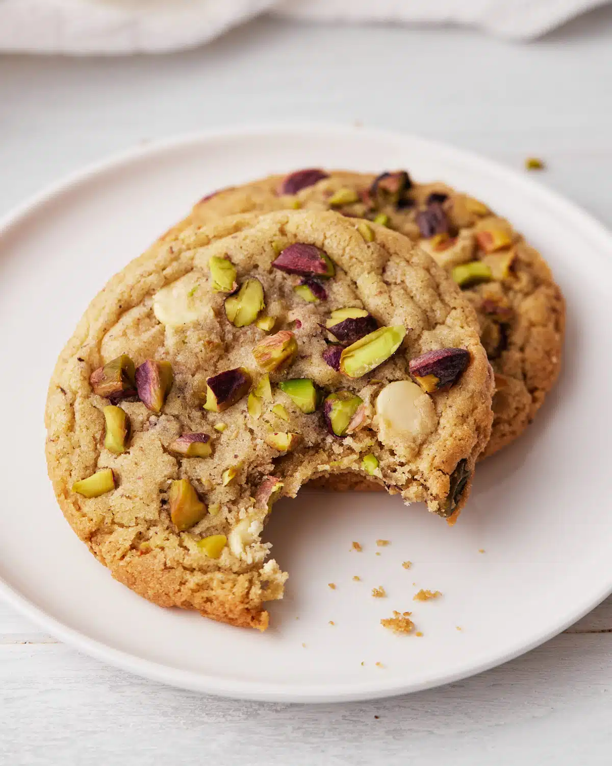 two pistachio cookies on a plate, one with a bite taken out.