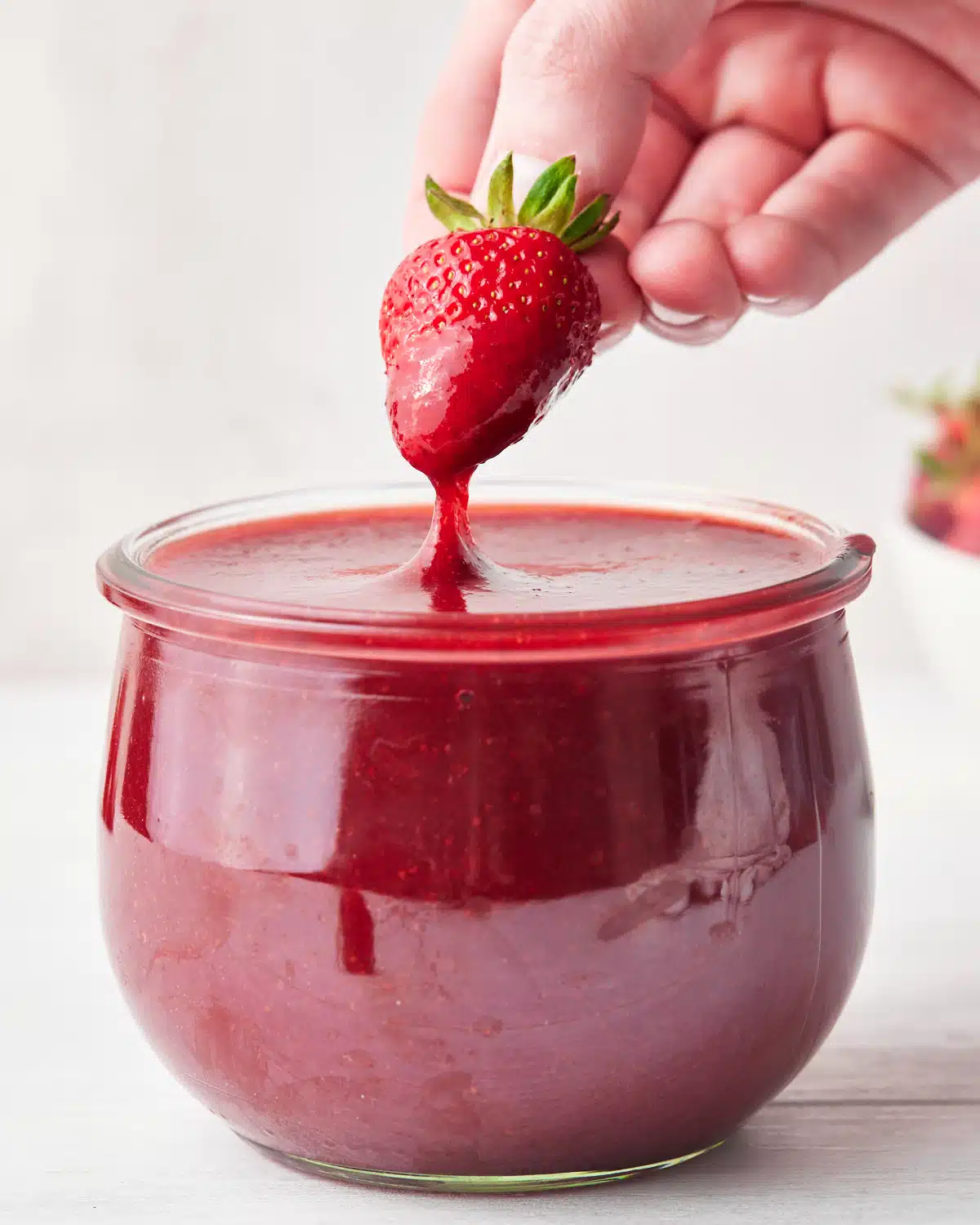 strawberry puree (strawberry coulis) in a glass jar, with a fresh strawberry being dipped in it. 