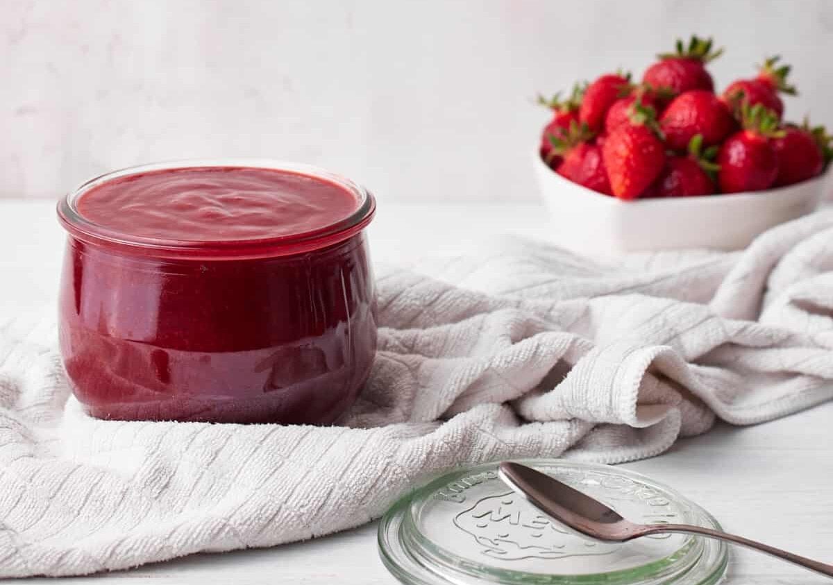 strawberry puree (strawberry coulis) in a glass jar with a bowl of fresh strawberries in the background.