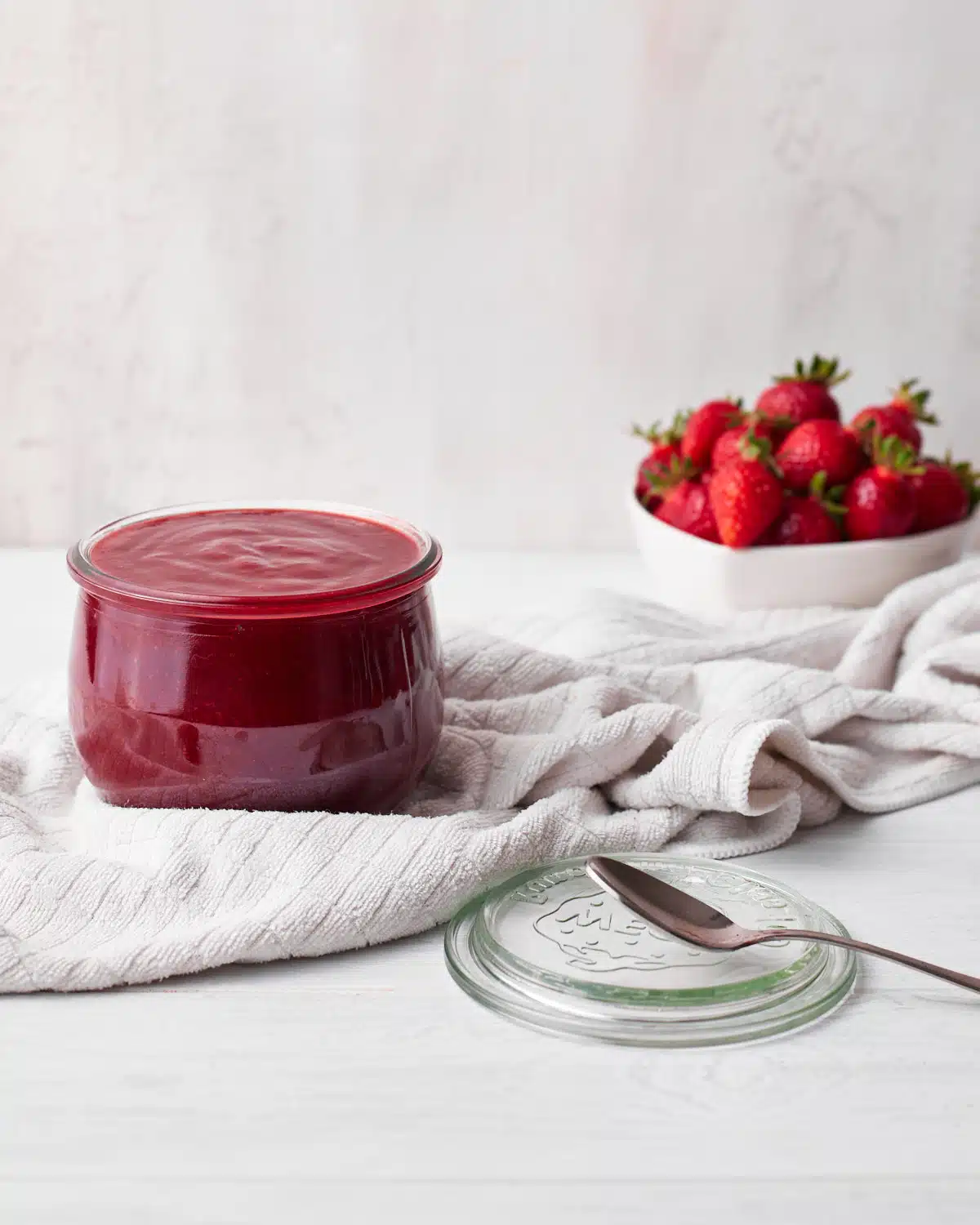 strawberry puree (strawberry coulis) in a glass jar with a bowl of fresh strawberries in the background. 