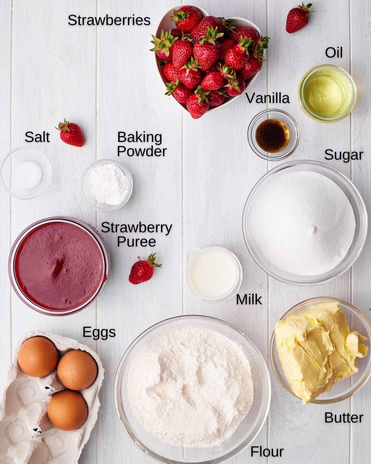 Labelled ingredients for strawberry cupcakes, including: strawberries, oil, vanilla, salt, baking powder, sugar, strawberry puree, milk, butter, eggs, and flour.
