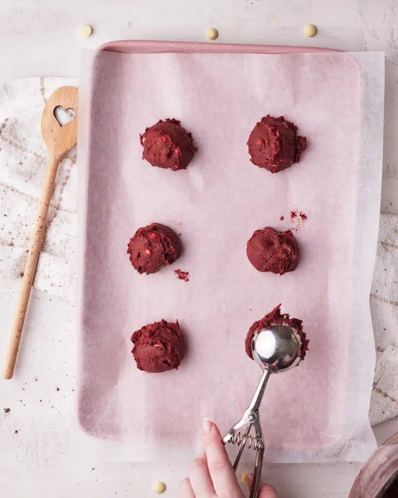 Scooping balls of red velvet cookie dough onto a baking sheet ready to be baked.