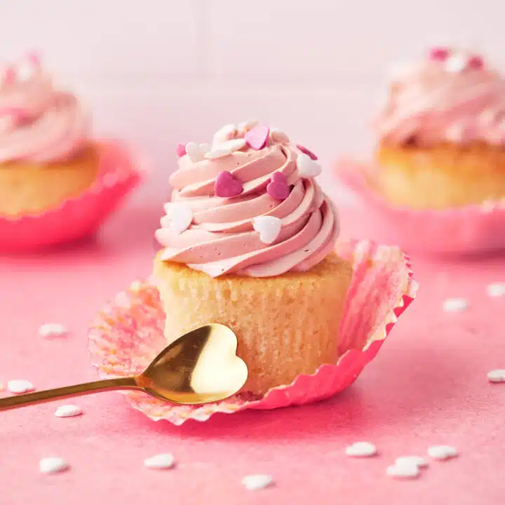 Valentines cupcakes on a pink background with white heart sprinkles all around.