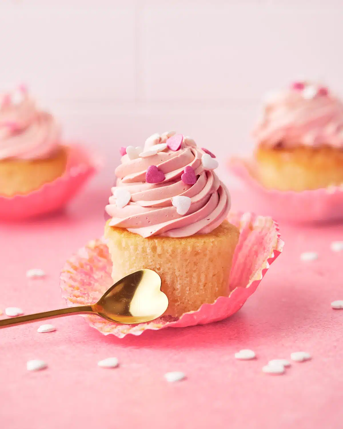 Valentines cupcakes on a pink background with white heart sprinkles all around.