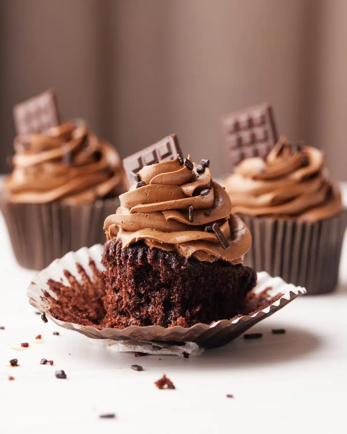 Three chocolate cupcakes on a table with chocolate buttercream frosting and a tiny chocolate bar sticking out the side.