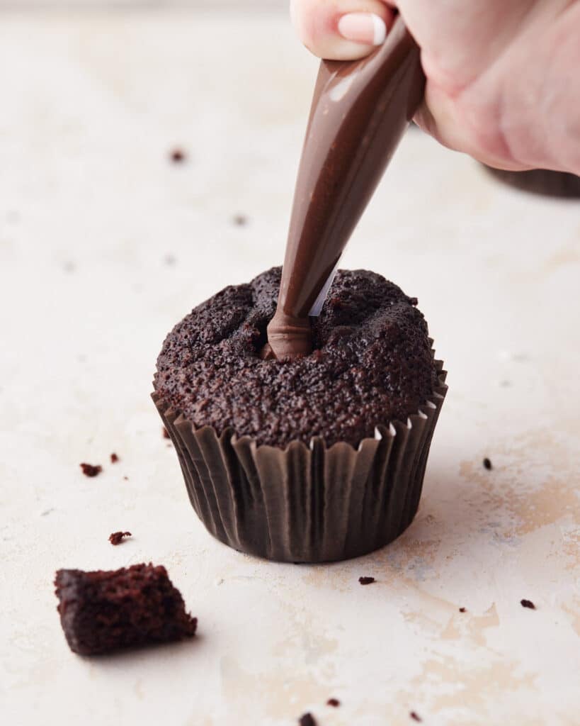 Chocolate cupcake with the center cored out, being filled with chocolate ganache through a piping bag.