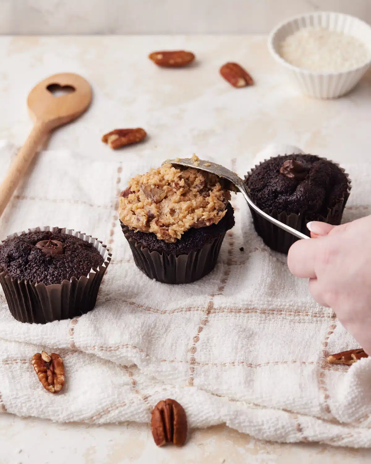 German chocolate cupcakes being decoraated with a spoon spreading the coconut pecan frosting on top of the cupcake. 