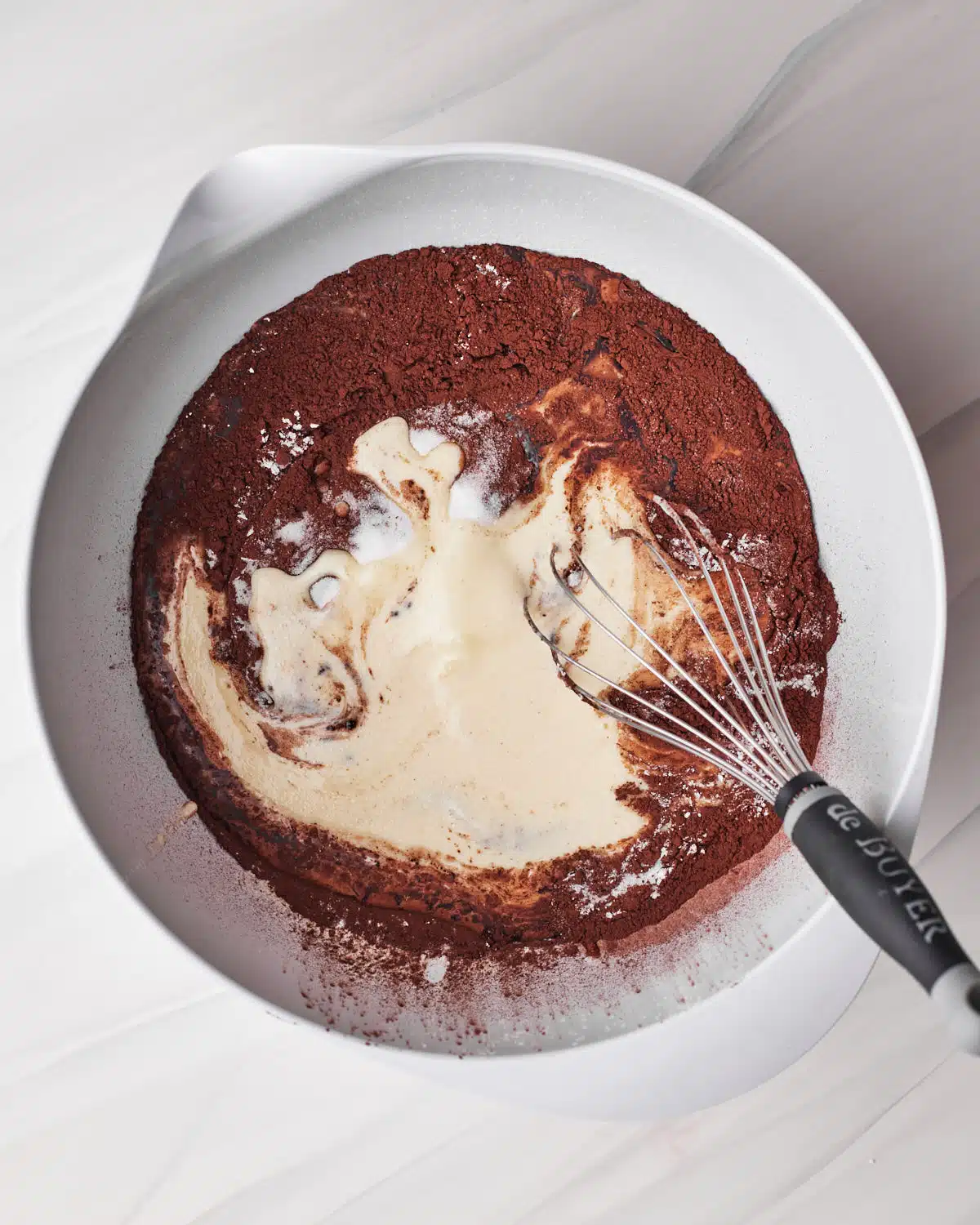 Wet ingredients and dry ingredients to make chocolate cupcakes in a large white bowl with a whisk.