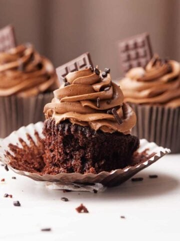 Three chocolate cupcakes on a table with chocolate buttercream frosting and a tiny chocolate bar sticking out the side.