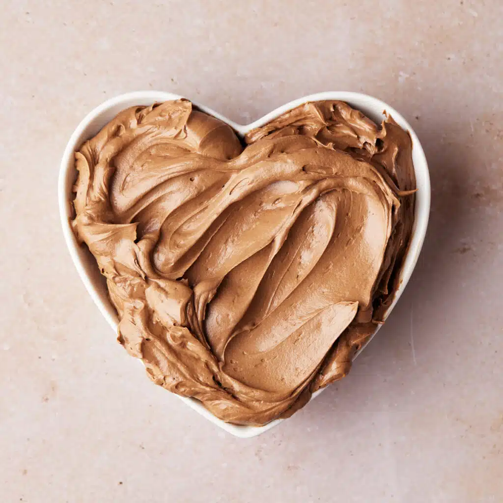 Silky smooth chocolate buttercream frosting in a heart shaped bowl, with ripples drawn in it to show how smooth it is.