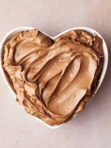 Silky smooth chocolate buttercream frosting in a heart shaped bowl, with ripples drawn in it to show how smooth it is.