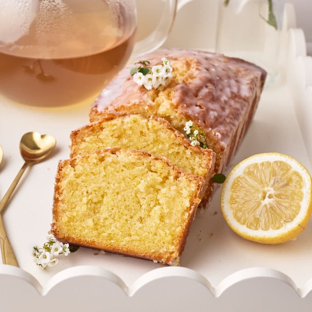 Lemon Drizzle Cake on a white serving tray with two slices sloping against the side, a fresh slice of lemon next to it.