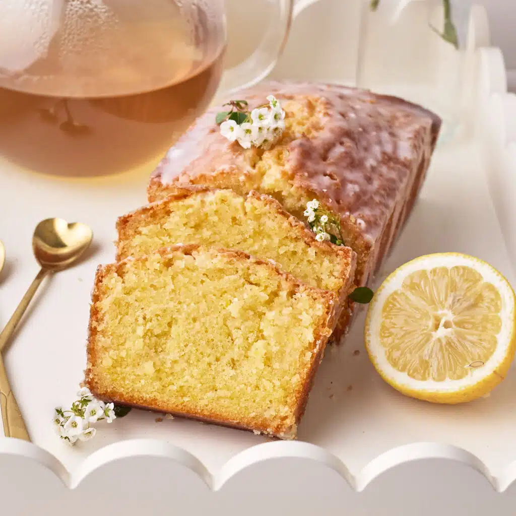Lemon Drizzle Cake on a white serving tray with two slices sloping against the side, a fresh slice of lemon next to it.
