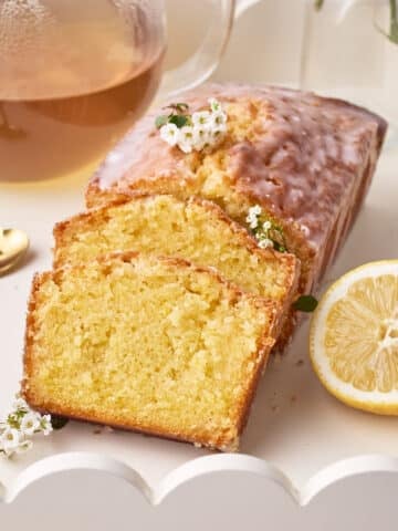 Lemon Drizzle Cake on a white serving tray with two slices sloping against the side, a fresh slice of lemon next to it.