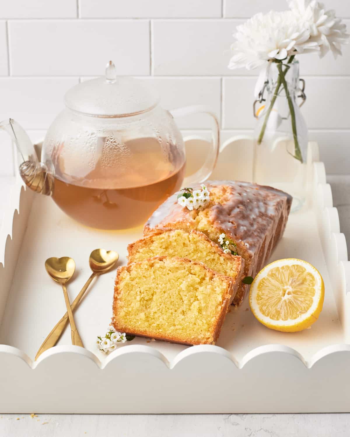 Lemon Drizzle Cake on a white serving tray with fresh white flowers, a glass teapot filled with tea, a slice of fresh lemon, and two heart shaped spoons. 