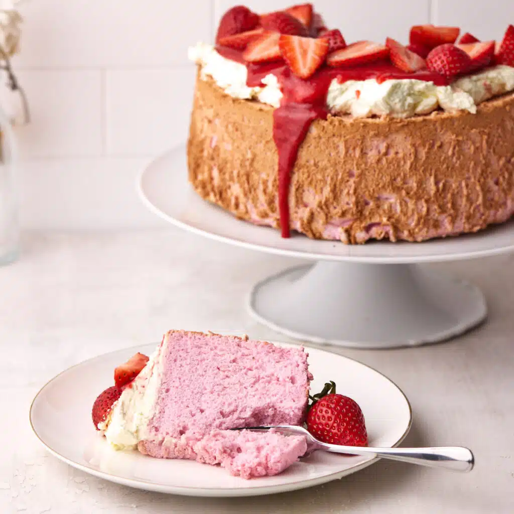 Pink Strawberry angel food cake with a slice taken out, the slice is sitting on a place in front with fresh strawberries.