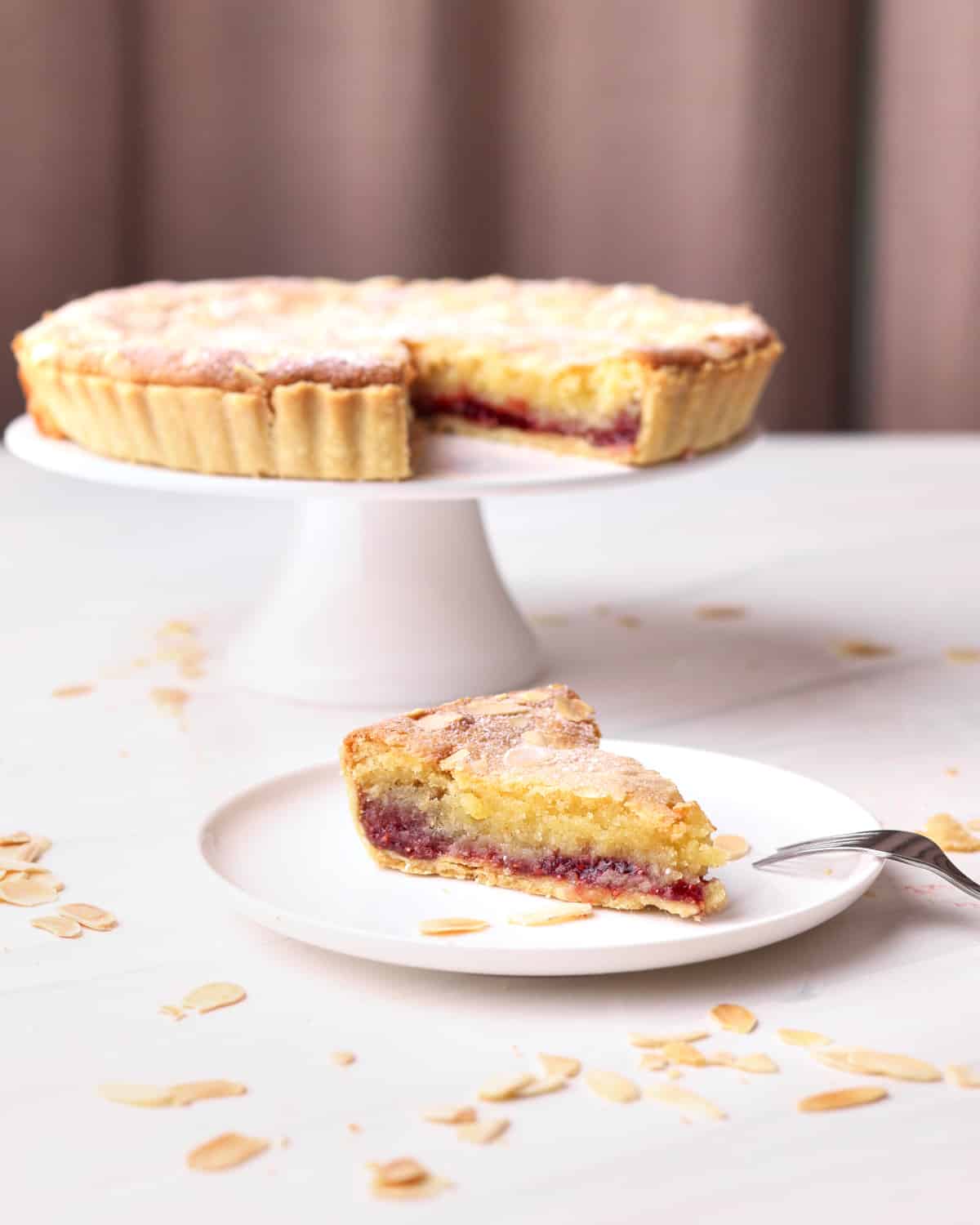 Bakewell tart with a slice taken out of it, the slice is on a plate in front of the tart. 