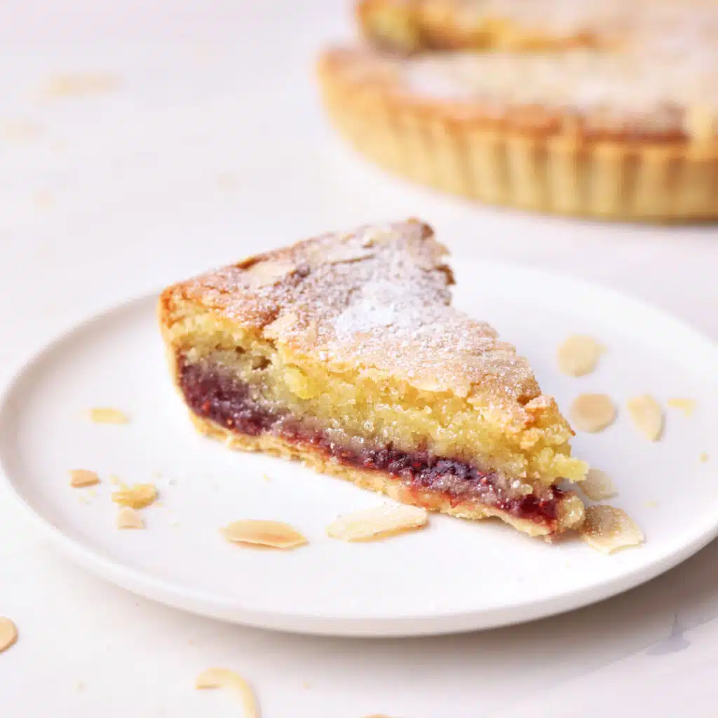 A slice of Bakewell tart, sitting on a plate with slivered almonds scattered around.